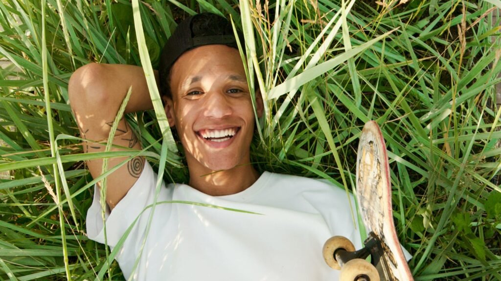 Young man lying in the grass on a sunny day, smiling with a skateboard beside him.