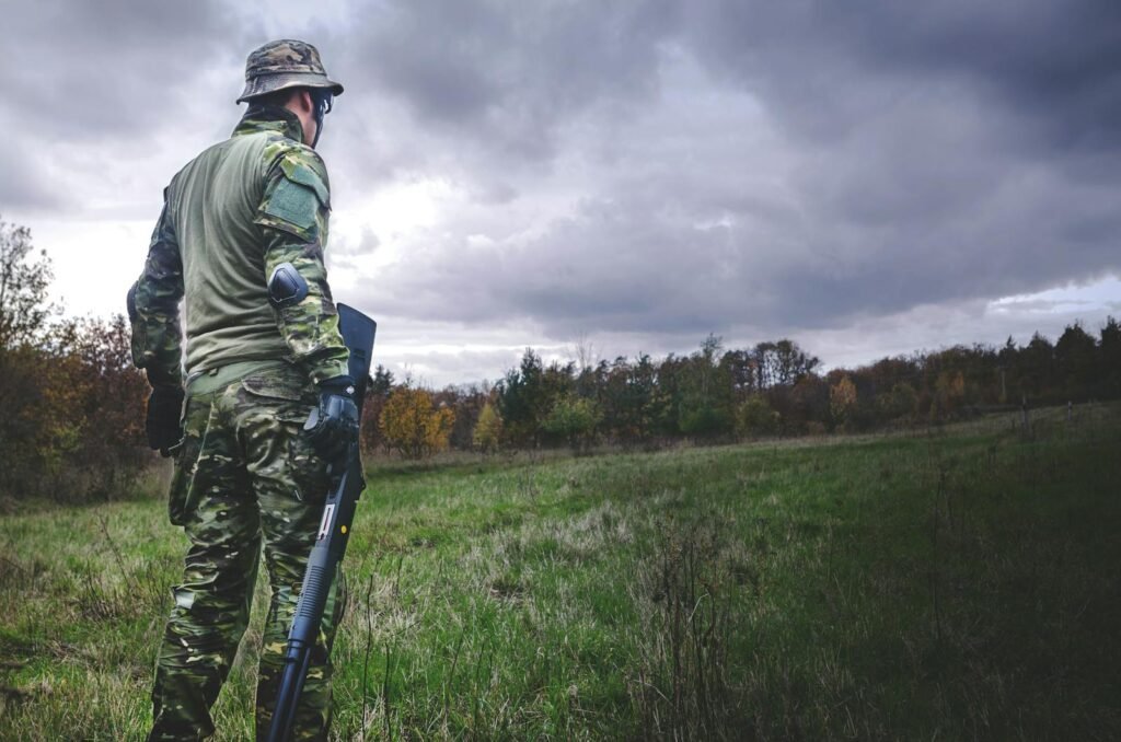 Soldier in camouflage with a rifle stands in a grassy field under a cloudy sky.
