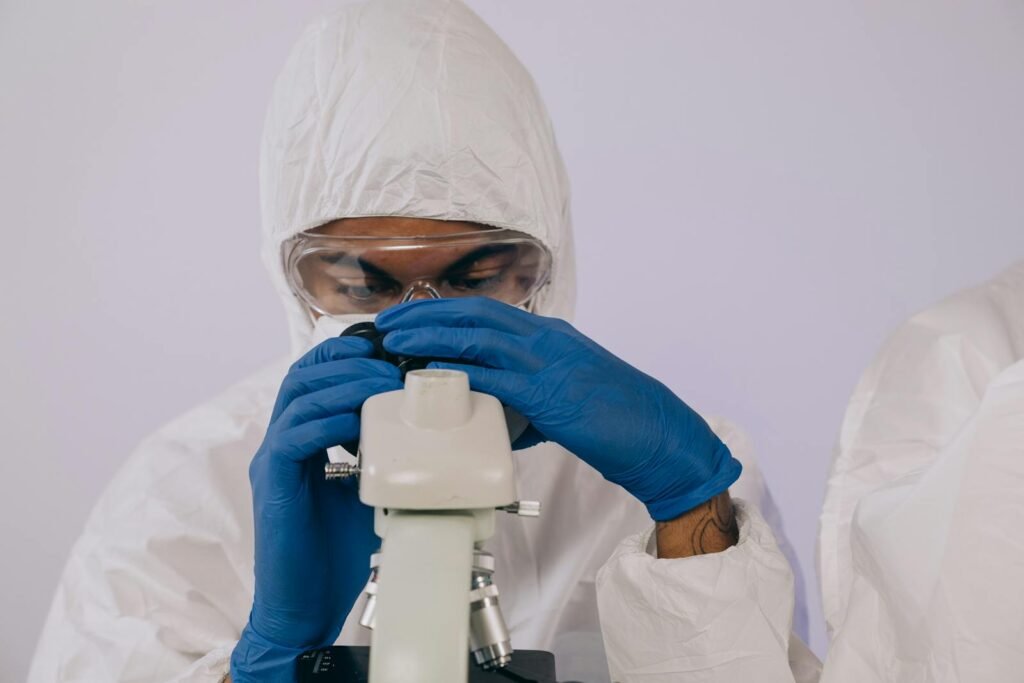 Microbiologist in protective gear examines samples through a microscope in a lab setting.