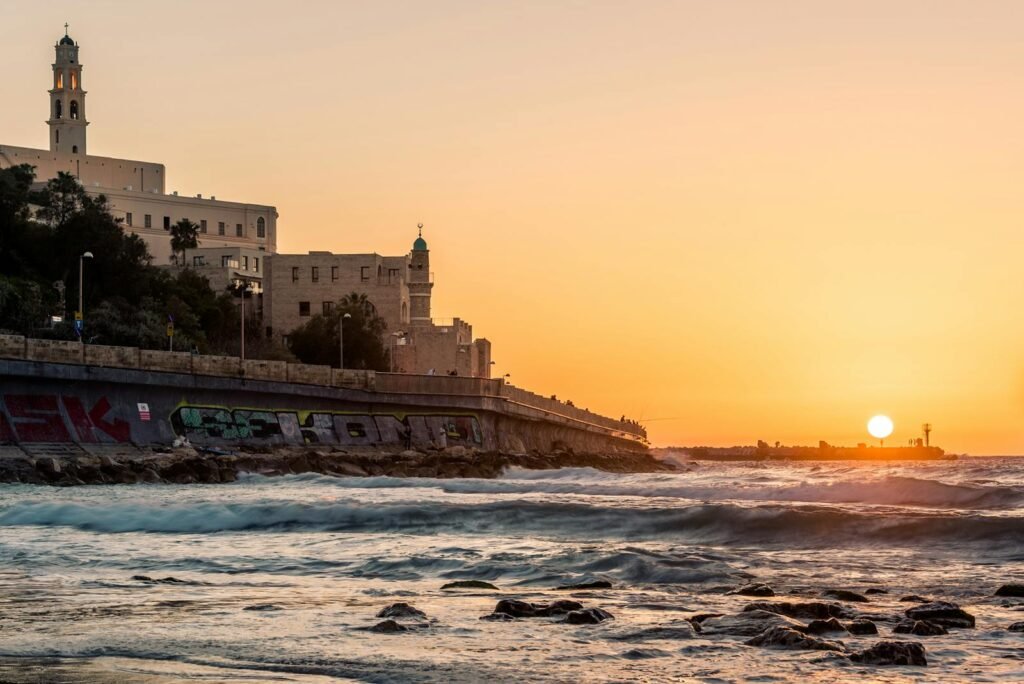 Sunset view of a historic coastal cityscape with buildings and ocean waves.