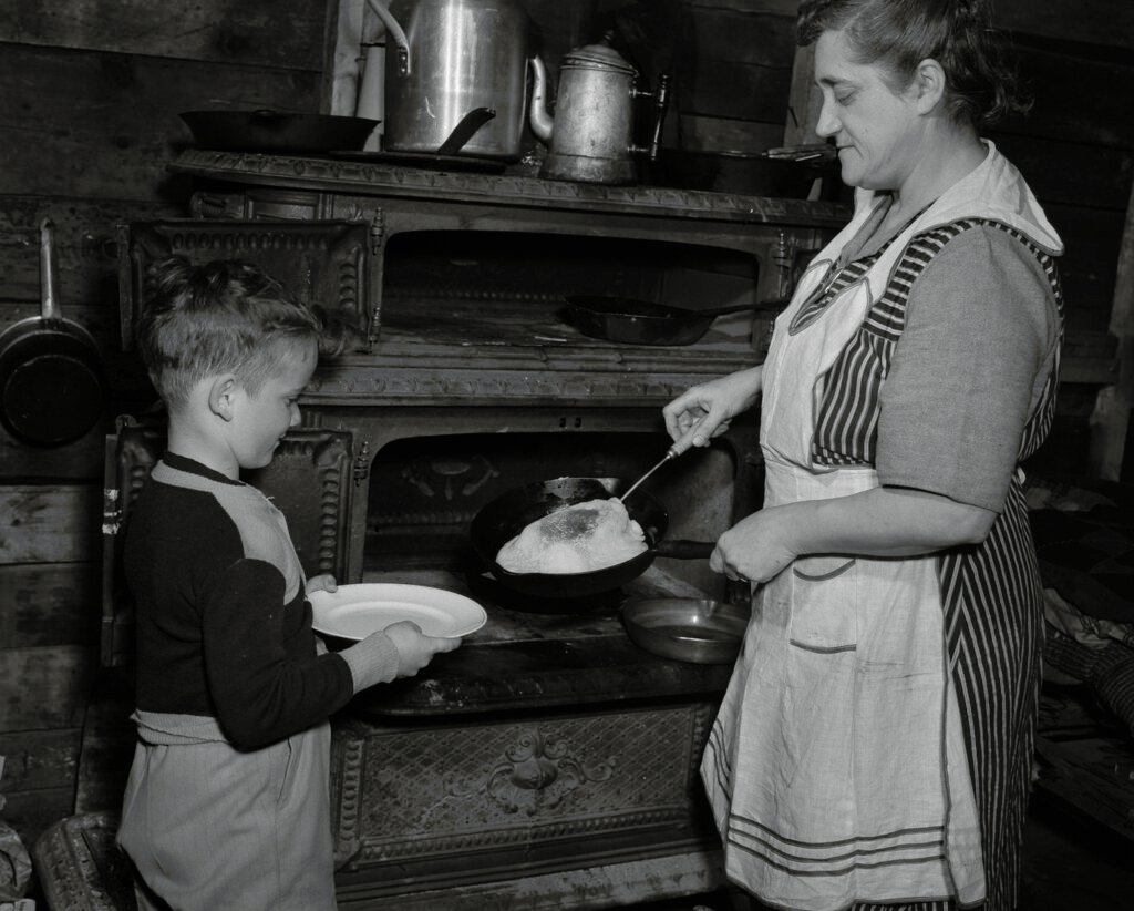 Child and mother cooking pancakes on two bridge stove - sugar bush camp / Un enfant et sa mère font cuire des crêpes dans un four à deux étages d’une érablière