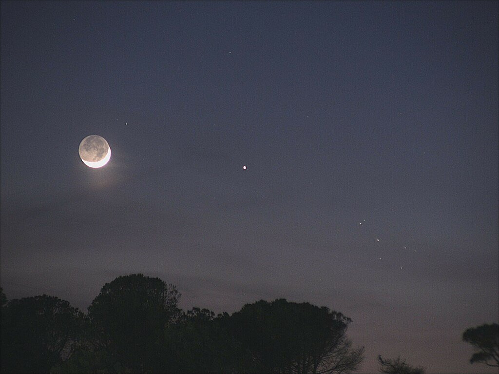 Image title: Moon-Mercury-Pleiades ConjunctionAuthor: Giulio ColomboCountry: Italy This photograph shows the young lunar crescent, some of the nine brightest stars of the Pleiades (with one behind a cloud) on the right, and the planet Mercury, looking slightly red, in the middle of the image. This picture is rather suggestive of the idea that the Pleiades might possibly consist of seven stars. However, the viewer is misled by the clouds; five of the stars form a tiny chariot, one is next to the handle, and three are at the other end of the quadrilateral. Eight stars would be clearly visible if there were no clouds. This configuration of the young Moon next to the Pleiades is visible only in the northern hemisphere spring. Thus it was used by the ancient Babylonians to determine the second month of their year and to judge whether or not an intercalary month was necessary. At least as early as the second millennium before the common era, the Babylonians used several asterisms for each month, with another one of them reappearing every five days after invisibility during daylight. To determine the necessity of intercalation in order to synchronise the solar and the lunar year, the Babylonians used several asterisms, not only the Pleiades. For instance, they also made use of the bright stars Arcturus and Sirius, and they observed a configuration with the Moon as well as heliacal phenomena. The modern Jewish and modern Islamic traditions still make use of some of the Babylonian astronomical rules. However, given that the constellations have shifted as a result of precession, and the fact that nowadays we also have computational means to calculate our calendars, this configuration of the small crescent Moon and the Pleiades is less useful, though it remains exceptionally beautiful. Thus the ancient Babylonian and middle Babylonian tradition survives only rudimentarily. Furthermore, it is unlikely that it is depicted in the Nebra Disc from Bronze Age Europe, as has long been claimed. This image was taken on Elba Island, Italy, in May 2022. Also see image in Zenodo: https://doi.org/10.5281/zenodo.7425575