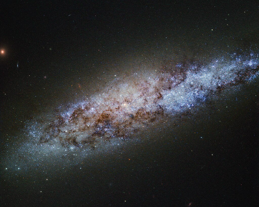 This bundle of bright stars and dark dust is a dwarf spiral galaxy known as NGC 4605, located around 16 million light-years away in the constellation of Ursa Major (The Great Bear). This galaxy’s spiral structure is not obvious from this image, but NGC 4605 is classified as an SBc type galaxy — meaning that it has sprawling, loosely wound arms and a bright bar of stars cutting through its centre. NGC 4605 is a member of the Messier 81 group of galaxies, a gathering of bright galaxies including its namesake Messier 81 (heic0710), and the well-known Messier 82 (heic0604a). Galaxy groups like this usually contain around 50 galaxies, all loosely bound together by gravity. This group is famous for its unusual members, many of which formed from collisions between galaxies. With its somewhat unusual form, NGC 4605 fits in well with the family of perturbed galaxies in the M81 group, although the origin of its abnormal features is not yet clear. The Messier 81 group is one of the nearest groups to our own, the Local Group, which houses the Milky Way and some of its well-known neighbours, including the Andromeda Galaxy and the Magellanic Clouds. Galaxy groups provide environments where galaxies can evolve through interactions like collisions and mergers. These galaxy groups are then lumped together into even larger gatherings of galaxies known as clusters and superclusters. The Local and Messier 81 groups both belong to the Virgo Supercluster, a large and massive collection of some 100 galaxy groups and clusters. With so many galaxies swarming around, NGC 4605 may seem unremarkable. However, astronomers are using this galaxy to test our knowledge of stellar evolution. The newly-formed stars in NGC 4605 are being used to investigate how interactions between galaxies affect the formation, evolution, and behaviour of the stars within, how bright stellar nurseries come together to form stellar clusters and stellar associations, and how these stars evolve over time. And that's not all — NGC 4605 is also proving to be a good testing ground for dark matter. Our theories on this hypothetical type of matter have had good success at describing how the Universe looks and behaves on a large scale — for example at the galaxy supercluster level — but when looking at individual galaxies, they have run into problems. Observations of NGC 4605 show that the way in which dark matter is spread throughout its halo is not quite as these models predict. While intriguing, observations in this area are still inconclusive, leaving astronomers to ponder over the contents of the Universe.