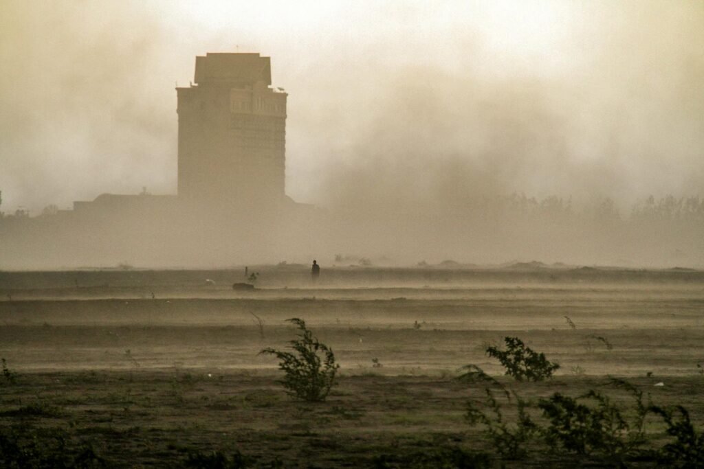 A dramatic scene of a dust storm engulfing a building and sparse vegetation in the foreground.