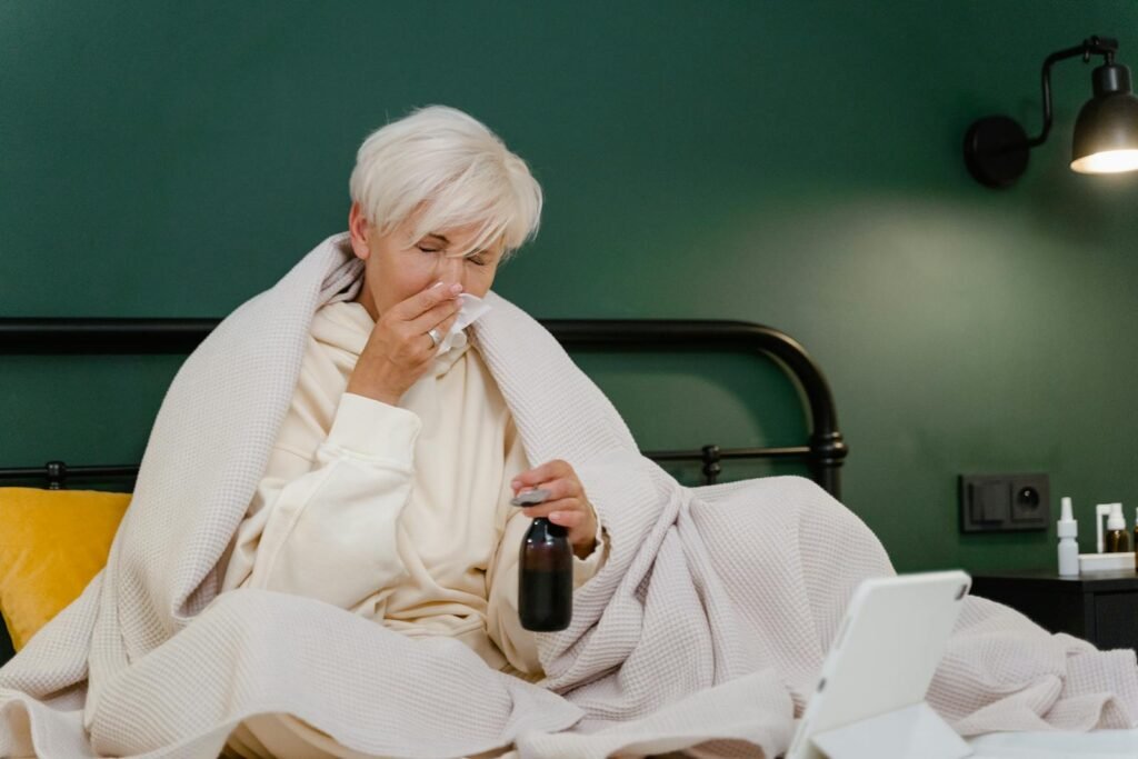 Elderly woman sits in bed feeling unwell, covered with a blanket, holding medicine.