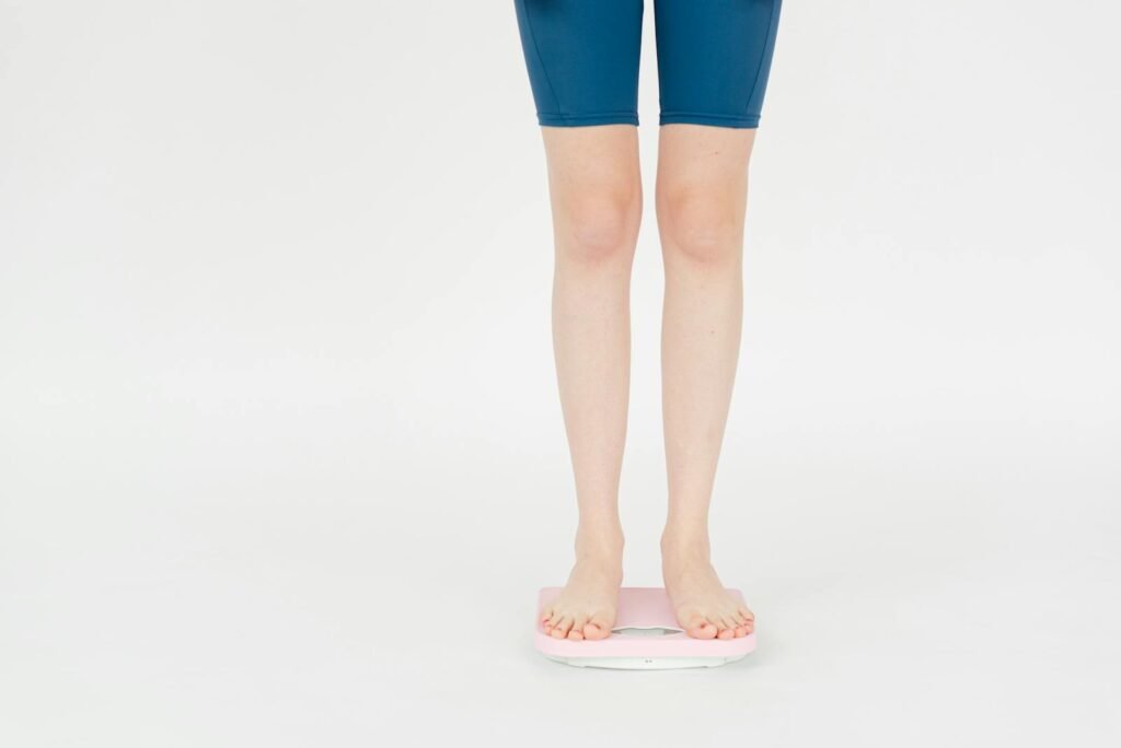 Crop anonymous female in sportswear measuring weight on scales for body control against white background