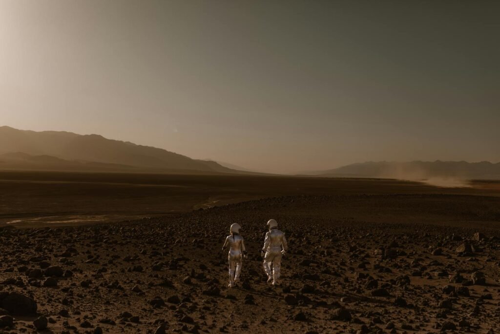 Two astronauts in spacesuits walk across a Mars-like desert landscape during the day.