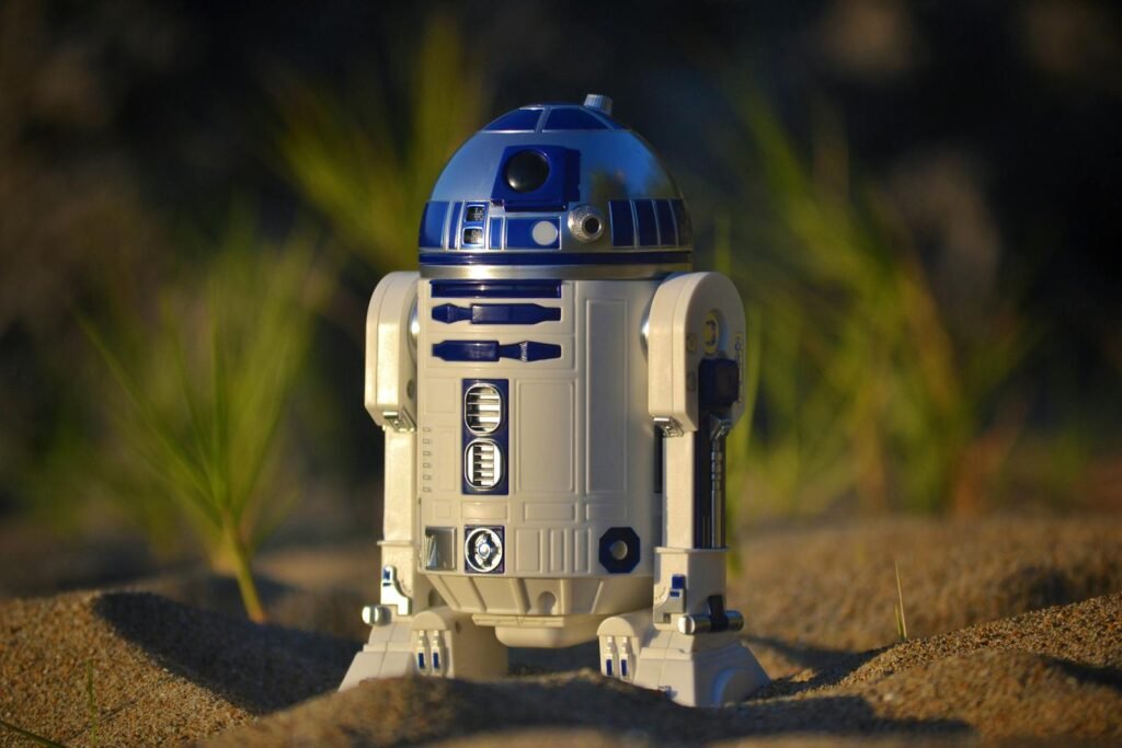 Close-up of an R2-D2 toy standing on sand, set against a green background in bright daylight.
