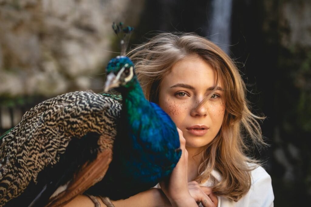 A woman with freckles poses outdoors with a peacock in a serene natural scene.