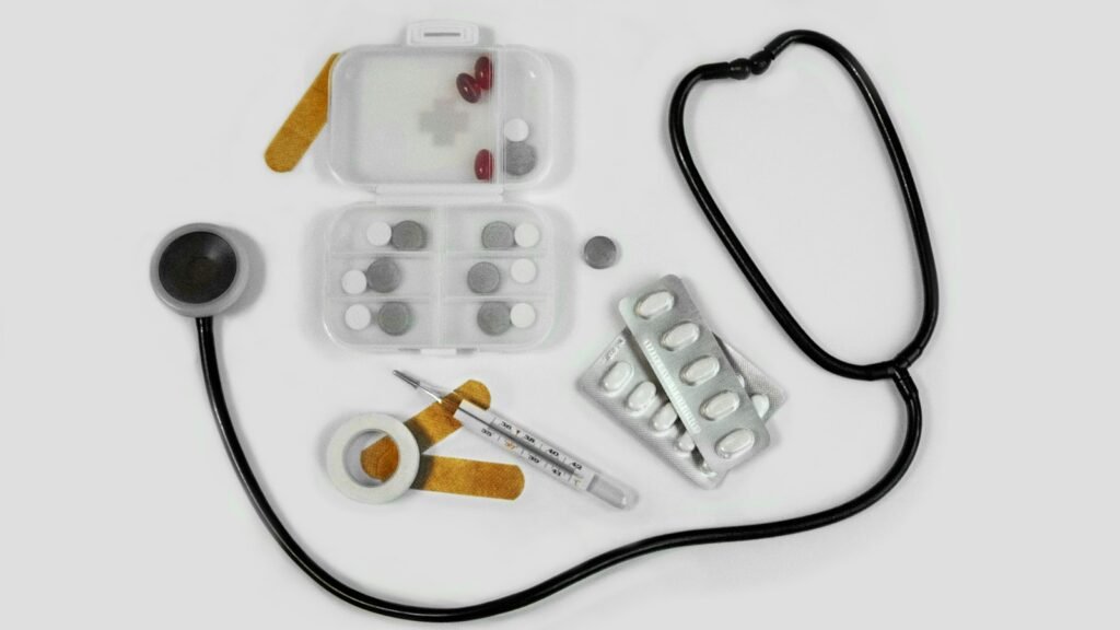 Basic medical kit with a stethoscope, blister packs of pills, a thermometer, and adhesive tape.